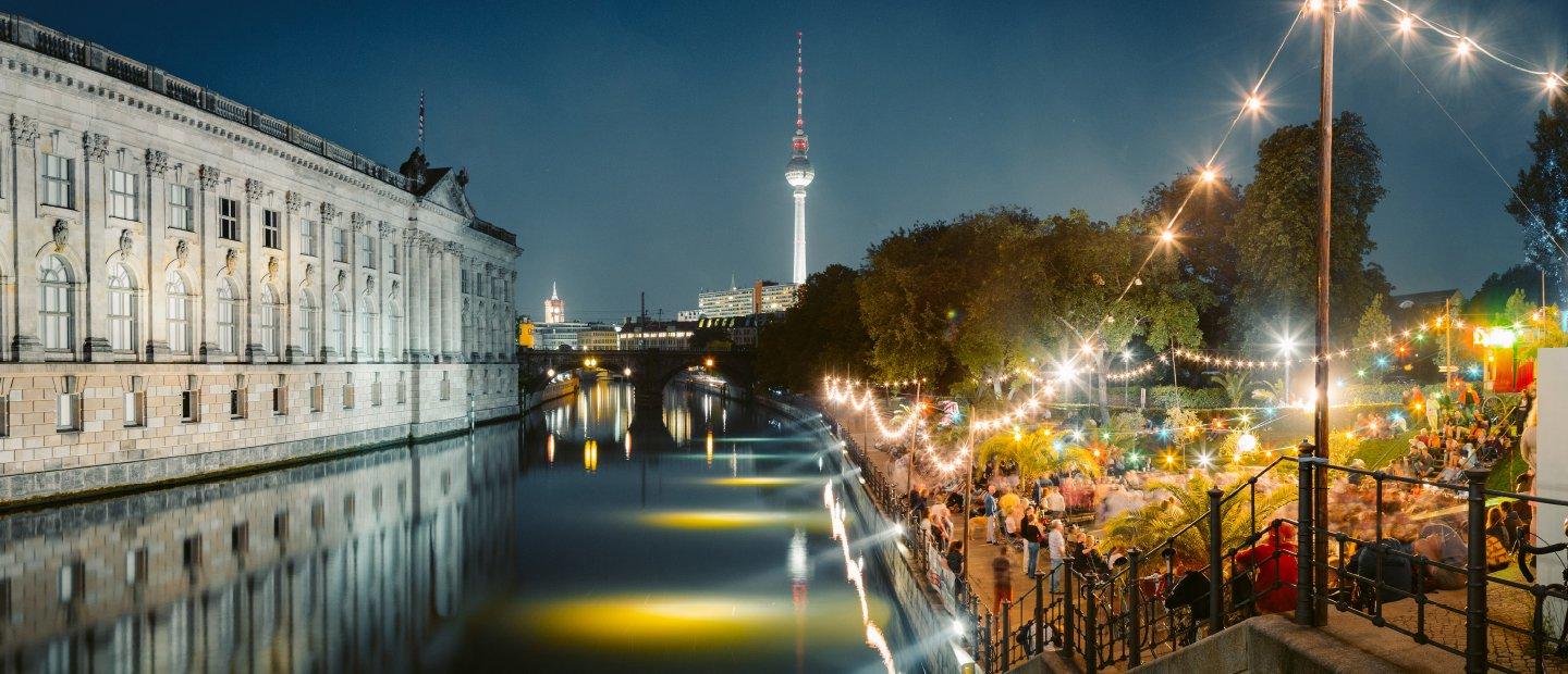 A canal in Germany with a large building on one side and a brightly lit patio on the other.